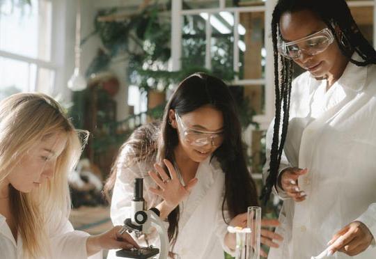 stock photo of a black woman with braids, an asian woman and a blonde woman wearing clear safety goggles and white lab coats huddled around various lab equipment 