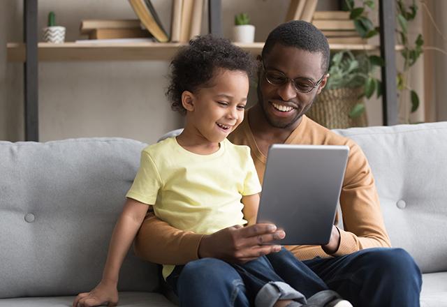 兴奋的非裔美国爸爸拿着平板电脑和可爱的学龄前儿子坐在沙发上一起看卡通片, 微笑着快乐的黑爸爸和小男孩在家里的沙发上放松地玩游戏